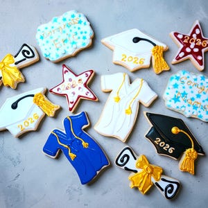 May include: Assortment of graduation-themed cookies, including mortarboards, diplomas, and stars. The cookies are decorated with white, blue, red, and gold icing, with the year 2026 and congratulatory messages.