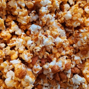 May include: Close-up of caramel popcorn with a mix of white and golden-brown kernels. Some pieces are coated in a shiny, amber-colored glaze, and some have peanuts mixed in.