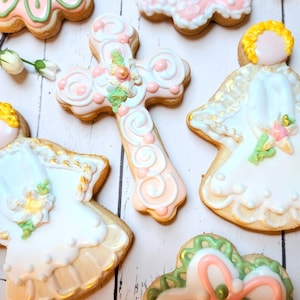 May include: Assortment of decorated Easter cookies, including angel-shaped cookies with yellow hair and white dresses, a cross-shaped cookie, and other floral-themed cookies. The cookies are decorated with pink, white, and green icing.