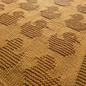 May include: A close-up of a knitted blanket with a mustard yellow color and a repeating pattern of hearts.