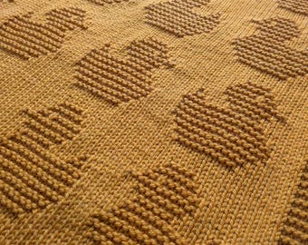 May include: A close-up of a knitted blanket with a mustard yellow color and a repeating pattern of hearts.