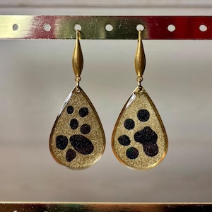 Puede incluir: Pendientes dorados en forma de lágrima con un fondo dorado texturizado y formas abstractas negras. Los pendientes se muestran en un soporte dorado. Los pendientes tienen una longitud de unos 5 cm.