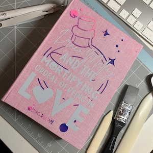 May include: A pink fabric-covered journal with the title "Draco Malfoy and the Mortifying Ordeal of Being in Love" in white lettering. The journal has a purple and pink illustration of a bottle with a star design.