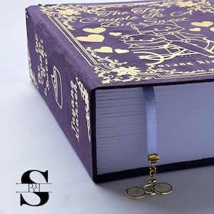 May include: A purple journal with gold embossed text and illustrations, including hearts and a couple in a carriage. The journal has a light blue ribbon bookmark with a gold bicycle charm. The text on the cover reads "The Top Is A Couple Sweet Bit."