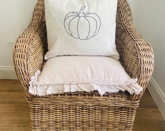 May include: A square off-white throw pillow featuring a black embroidered pumpkin outline centered on the front. Below it sits a rectangular light beige pillow with a decorative ruffled fringe edge, both resting on a woven brown wicker armchair.