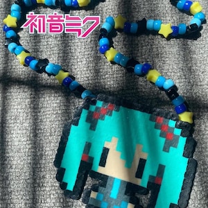 Può includere: Una collana di perline fatta a mano con un ciondolo a forma di personaggio anime pixelato. La collana ha una catena di perline blu, gialle e nere con perline a forma di stella. Il ciondolo è turchese, nero, rosso e beige. Il testo giapponese "Hatsune Miku" è visibile.