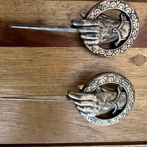 Pode incluir: Dois broches de metal dourado em forma de mão com um design circular. Cada broche tem uma mão com uma adaga presa ao pulso.