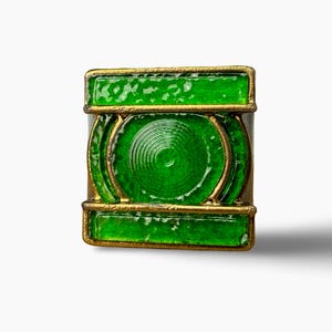 May include: A square-shaped ring featuring a vibrant green enamel inlay set within a metallic gold-toned frame. The design includes a central circular motif with concentric ridged detailing, flanked by horizontal rectangular green panels.