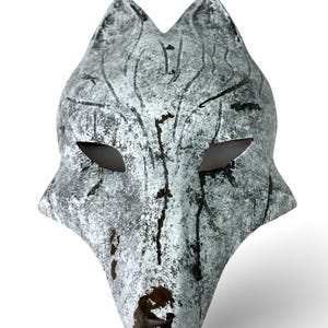 Könnte beinhalten: Eine handgefertigte Wolfsmaske mit einem verwitterten, strukturierten Finish. Die Maske ist überwiegend grau mit weißen Akzenten und dunkelbraunen Akzenten, mit Augenausschnitten und detaillierten Gesichtszügen. Für Kostüm- oder Dekorationszwecke.