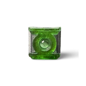 Puede incluir: Un logotipo verde de Green Lantern de vidrio. El logotipo tiene forma cuadrada con un emblema circular en el centro. El vidrio tiene una calidad reflectante y los bordes están delineados en un color más oscuro.