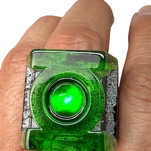 May include: A square-shaped ring featuring a translucent, vibrant green center with the circular Green Lantern symbol embossed in the middle. The ring has a textured, metallic silver-colored base that contrasts with the bright green top surface.