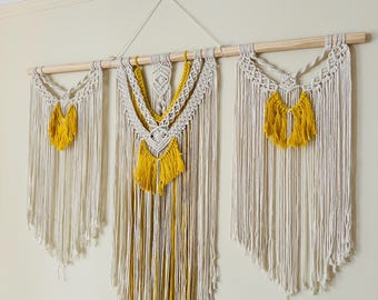 Pode incluir: Um conjunto de três tapeçarias de macramé de parede com uma haste de madeira. As tapeçarias são feitas com cordão de algodão de cor natural e têm borlas amarelas.