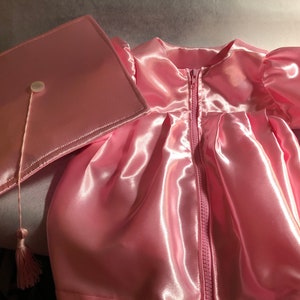 May include: A pink satin graduation gown with a matching pink satin cap and tassel. The gown has a zipper closure and the cap has a button on top.