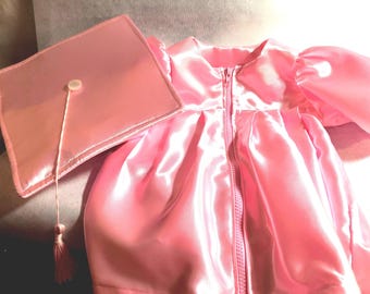 May include: A shiny pink graduation gown featuring a front zipper closure and puffed sleeves, accompanied by a matching square graduation cap with a pink tassel attached to a central button. The set is made of a smooth, reflective satin-like fabric.