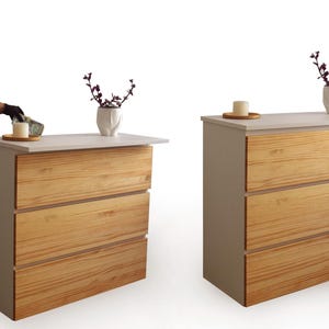 Könnte beinhalten: Zwei Holzkommoden mit je drei Schubladen. Die Schubladen haben eine helle Holzmaserung. Die Oberseiten sind weiß, mit einer kleinen Vase mit lila Stielen und einer Kerze auf einem Holzuntersetzer. Höhe ca. 80 cm.