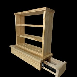 Puede incluir: Una estantería de madera de color claro con tres estantes y un cajón. La unidad tiene forma rectangular y está hecha de madera sin terminar. El cajón está abierto, revelando un riel de metal negro. Los estantes tienen múltiples agujeros.
