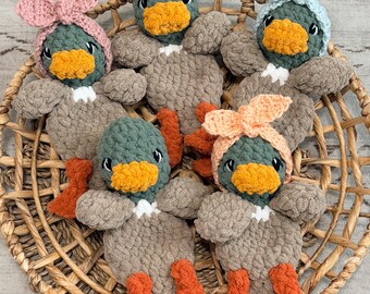 May include: Five handmade crochet duck plushies with dark green heads, orange beaks and feet, and light brown bodies. Three of the ducks wear crocheted headbands in light pink, light blue, and peach colors, all arranged inside a round woven basket.
