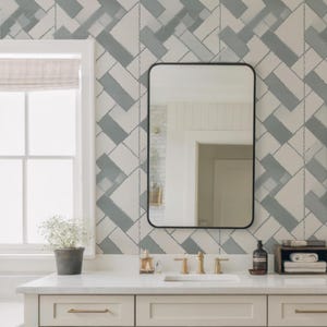 May include: A bathroom scene featuring a rectangular black-framed mirror above a white vanity. The vanity has gold fixtures and a white countertop. The wall is decorated with a geometric tile pattern in shades of gray and white. A window with a white frame is visible.