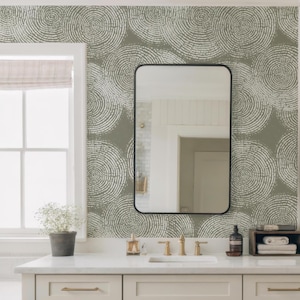 May include: A bathroom vanity with a white countertop, a rectangular black-framed mirror, and a patterned wallpaper. The vanity has light-colored cabinets with gold-toned hardware. A potted plant and toiletries are visible.