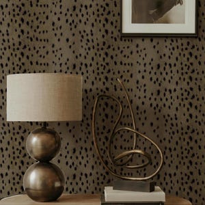 Könnte beinhalten: Eine bronzefarbene Tischlampe mit kugelförmigem Sockel und beigem Lampenschirm steht auf einem Holztisch. Eine goldfarbene abstrakte Skulptur und gerahmte Schwarz-Weiß-Kunst befinden sich ebenfalls auf dem Tisch. Der Hintergrund ist eine braune Wand mit schwarzen Punkten.