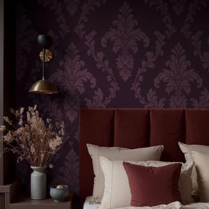 Può includere: Interno di una camera da letto con testiera bordeaux, cuscini color crema e una coperta rosso scuro. La parete è decorata con carta da parati damascata viola scuro. Un comodino contiene un vaso con fiori secchi e una piccola ciotola. Un'applique in ottone fornisce l'illuminazione.