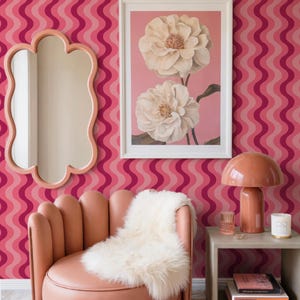 May include: A room with a pink and red wavy patterned wallpaper. A pink scalloped mirror hangs on the wall. A framed floral print and a pink mushroom lamp sit on a side table next to a pink chair with a white fur throw.