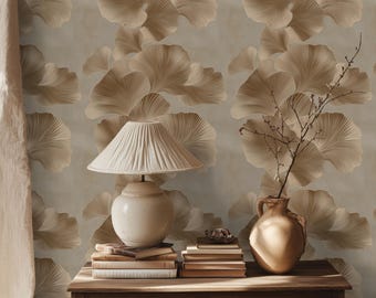 Op de afbeelding: Een beige lamp met een witte kap staat op een houten tafel met een stapel boeken en een bruine vaas met een droge tak. De muur achter de tafel is bedekt met behang met een patroon van bruine ginkgo bladeren.
