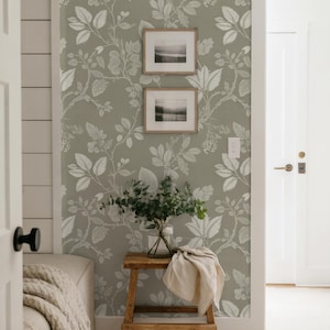 May include: A room with sage green floral wallpaper and two framed landscape prints. A small wooden stool holds a vase of greenery and a folded beige cloth. The room has white trim and a partially visible bed with a cream-colored blanket.