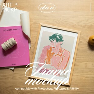 May include: A framed art print mockup featuring a portrait with pink and green hues. The image is in a light wood frame, with a pink book titled "JAPANESE PRINTS" and a roll of twine. The words "Frame mockup" are displayed in a script font.