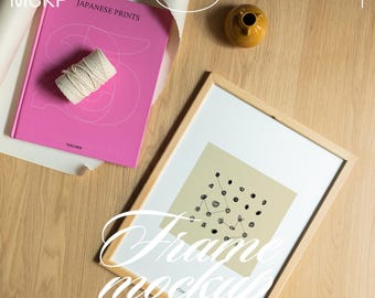 May include: A wooden framed art print with a black and white abstract design, a pink book titled "Japanese Prints", a small yellow vase, and a roll of twine on a wooden surface. The image also includes the text "Frame mockup".