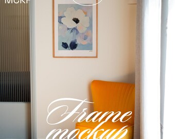 Op de afbeelding: Een ingelijste bloemenkunstprint met een witte bloem en blauwe accenten hangt aan een witte muur. Een oranje fluwelen stoel staat onder het kunstwerk. De afbeelding bevat de tekst "ART MCKP" en "Frame mockup".