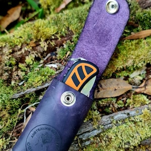 May include: A purple leather knife sheath with a black and orange folding knife inside. The sheath has a logo that says "Australian Bushcraft Co."