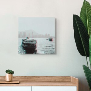 Könnte beinhalten: Leinwanddruck mit 3D-Effekt von zwei Booten auf dem Wasser. Die Boote sind dunkel mit weißen Akzenten, vor dem Hintergrund einer Stadtkulisse und Bergen unter einem dunstigen Himmel. Der Druck ist an einer weißen Wand befestigt.