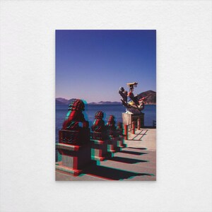Könnte beinhalten: Ein Foto mit 3D-Effekt, das rote und schwarze Statuen auf einem Pier zeigt, mit blauem Himmel, Meer und Bergen im Hintergrund. Eine größere, verzierte Skulptur ist zu sehen. Das Bild weckt ein Gefühl von Reise und kulturellem Erbe.