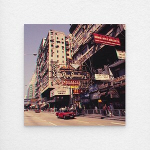 Könnte beinhalten: Eine Straßenszene in Hongkong mit einem roten Taxi auf einer Straße, die von hohen Gebäuden gesäumt ist. Schilder für "Roys Jewellery" und "Oriental Arts & Jewellery Co" sind zu sehen.