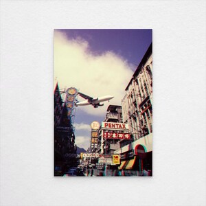 Könnte beinhalten: Ein Print im Vintage-Stil mit einer Stadtlandschaft und einem tief fliegenden Flugzeug. Das Bild hat einen 3D-Effekt mit Rot- und Blau-Farbtrennung. Gebäude mit Schildern und ein bewölkter Himmel sind sichtbar.
