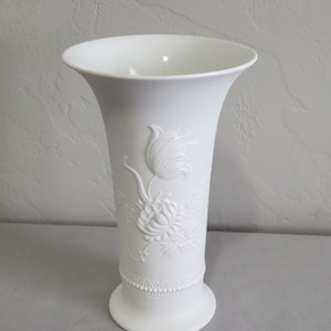 May include: A matte white porcelain vase featuring a flared rim and a tapered body. The exterior is decorated with a raised relief pattern of a blooming flower and foliage, complemented by a delicate beaded texture encircling the base.