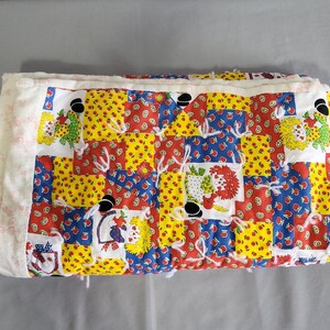 Puede incluir: Un edredón de retazos plegado con un diseño colorido. El edredón presenta cuadrados de tela roja, amarilla y azul con estampados florales y de frutas. Los bordes están acabados con un borde blanco y botones negros.