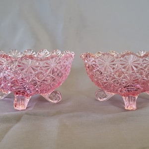 Könnte beinhalten: Zwei rosa Glasschalen mit Füßen und Sternmuster. Die Schalen haben einen gewellten Rand und werden von drei geschwungenen Beinen getragen. Das Glas hat ein strukturiertes, geschliffenes Aussehen.