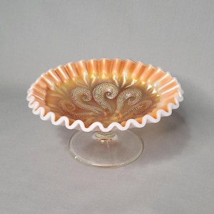 May include: An iridescent orange and gold glass pedestal bowl with a ruffled white rim. The bowl features a decorative pattern in the center and sits on a clear glass base. The bowl is likely vintage or antique.