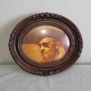 May include: Oval-shaped, brown framed portrait of a man wearing sunglasses and a hat. The frame has ornate detailing. The portrait is a sepia tone, with the man's face and hat in focus.