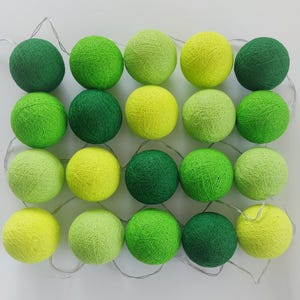 May include: A string of twenty spherical light covers arranged in a grid, featuring various shades of green and yellow thread. The spheres are connected by a thin, flexible silver wire, creating a decorative lighting element with a textured finish.
