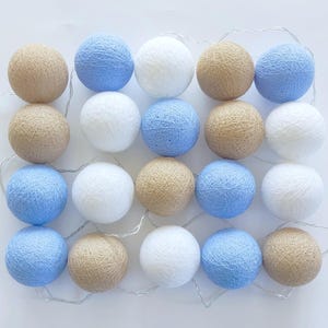 Può includere: Una stringa di luci decorative con palline di cotone di vari colori. Le luci sono disposte a griglia e includono palline azzurre, bianche e beige. Le luci sono infilate su un filo sottile e argentato, creando un bagliore soffuso e ambientale.