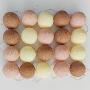 Op de afbeelding: Een reeks decoratieve katoenen bolletjes in verschillende kleuren, waaronder bruin, roze en crème. De lampjes zijn in een rasterpatroon gerangschikt en verbonden door een dunne, zilverkleurige draad. Perfect voor een warme sfeer.