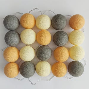 Può includere: Una stringa di luci decorative con venti sfere di cotone sferiche nei toni del grigio, giallo e arancione. Le luci sono disposte a griglia e collegate da un filo d'argento, creando un'atmosfera calda e accogliente.