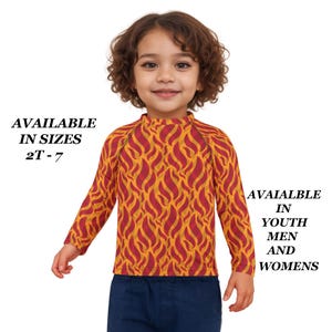 May include: A long-sleeved shirt with a flame pattern in shades of red and yellow. The shirt is worn by a child and is paired with blue pants. Text on the image reads "AVAILABLE IN SIZES 2T-7" and "AVAILABLE IN YOUTH MEN AND WOMENS."