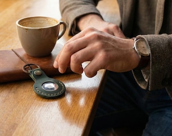 May include: A dark green leather keychain holder for an Apple AirTag, featuring a circular cutout, a metal snap button, and a bronze-toned keyring. The holder is constructed with visible stitching along the edges and secures a silver AirTag.