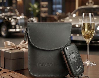 May include: A dark gray Faraday bag with a matching car key next to it. The bag has a curved flap and the text "FARADAYBAG" printed on the front. A glass of champagne and a gift box are also in the image.
