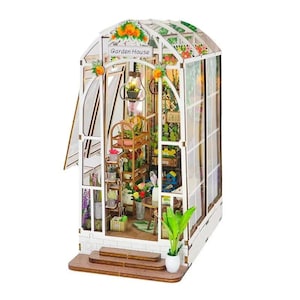May include: A miniature garden house model with a white frame, glass-like panels, and a sign reading Garden House. The interior features a wooden shelf with potted plants, a small table, a hanging light fixture, and a green plant in the front.