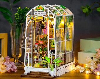 May include: A miniature wooden greenhouse model titled Garden House, featuring a white arched frame, clear panels, and interior shelves filled with tiny potted plants, flowers, and a hanging lantern, illuminated by warm internal LED lights.